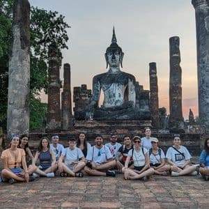 Eine WeRoad-Gruppenreise sitzt im Schneidersitz für ein Foto vor einer großen Buddha-Statue inmitten alter Tempelruinen bei Sonnenuntergang.