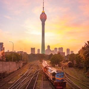 Un tren sobre vías férreas con una moderna torre en forma de loto y el horizonte urbano al fondo bajo un atardecer colorido.