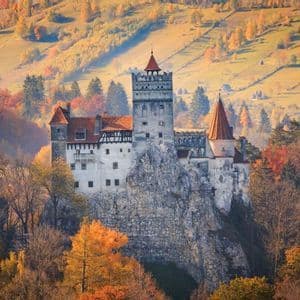 Eine mittelalterliche Burg mit roten Ziegeldächern, thronend auf einem felsigen Kliff, umgeben von dichtem Wald in herbstlichen Farben.