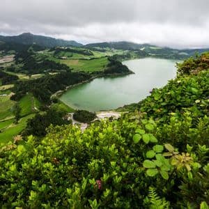 Vue plongeante sur un lac vert niché dans une vallée de collines verdoyantes et de champs sous un ciel nuageux.