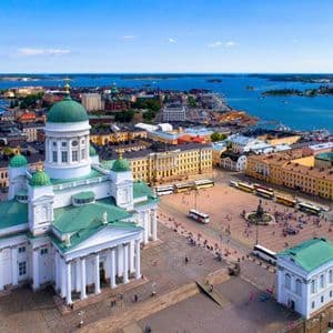 An aerial view of a large white cathedral with green domes overlooking a bustling city square and a bright blue sea with islands.