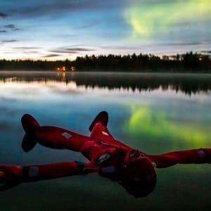 Una persona in tuta di sopravvivenza rossa galleggia supina in un lago tranquillo, con le verdi luci del nord che si riflettono sull'acqua al crepuscolo.