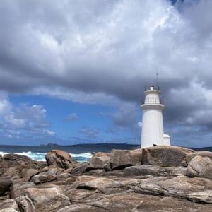 Un faro bianco si erge su una costa rocciosa accanto a un mare agitato sotto un cielo drammatico e nuvoloso.
