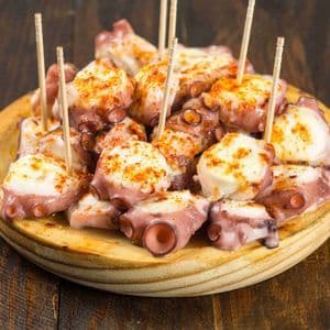 Un primo piano di pezzi di polpo cotti conditi con paprika, serviti con stuzzicadenti su un piatto di legno rotondo.