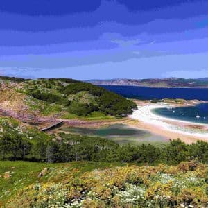Vista panorámica de una costa verde y rocosa con una playa de arena blanca y una bahía azul tranquila con veleros bajo un cielo despejado.