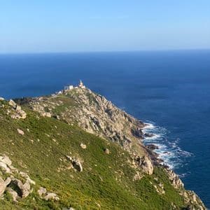 Un faro y edificios adyacentes se asientan en la punta de una península rocosa y verde, con vistas a un vasto océano azul bajo un cielo despejado.