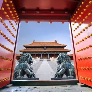 Large open red gates with golden knobs frame a traditional Chinese palace, guarded by two bronze lion statues.