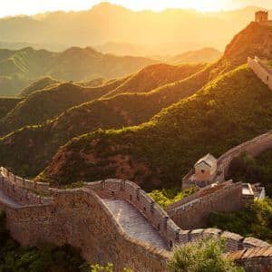 A long stone wall with watchtowers winds across lush green mountains under the golden light of a sunrise.