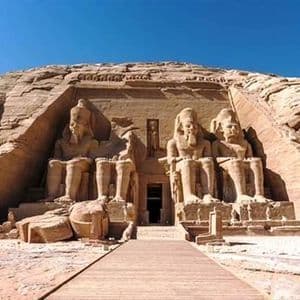 Una passerella di legno si avvicina all'ingresso di un grande tempio scavato nella roccia, affiancato da quattro colossali statue sedute.