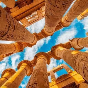 Una prospettiva dal basso di grandi colonne di pietra con geroglifici, che si innalzano verso un cielo azzurro con nuvole sparse.