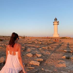 Una donna in abito bianco cammina attraverso un paesaggio roccioso verso un alto faro bianco durante un tramonto dorato.