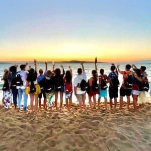 Un viaggio di gruppo WeRoad visto da dietro, in piedi su una spiaggia sabbiosa con le braccia alzate, mentre si guarda un tramonto sul mare.