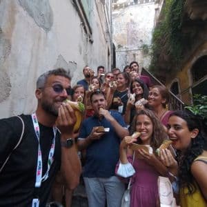 A WeRoad group trip smiles for the camera while eating cannoli on a narrow outdoor staircase.