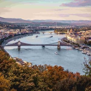 Ein erhöhter Blick auf eine Stadtlandschaft, die von einem breiten Fluss mit mehreren Brücken und Booten durchzogen wird, eingerahmt von Herbstlaub bei Sonnenuntergang.