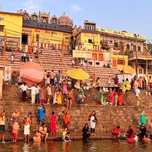 Una grande folla di persone in abiti colorati si raduna sui ghat di pietra, con molti che si bagnano nel fiume sotto tradizionali edifici gialli.