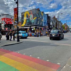 Un coin de rue en ville avec un passage piéton arc-en-ciel, des voitures, des piétons et des bâtiments recouverts de peintures murales colorées sous un ciel partiellement nuageux.