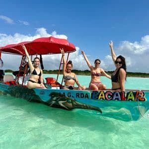 Vier Frauen in Bikinis auf einer WeRoad-Gruppenreise lächeln mit erhobenen Armen auf einem Boot in türkisfarbenem Wasser unter blauem Himmel.
