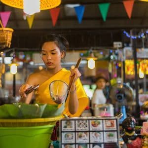 Eine Frau in traditioneller gelber Kleidung bereitet an einem hellen Marktstand Speisen mit Sieb und Zange zu.
