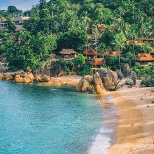 Luftaufnahme einer sandigen Strandbucht mit türkisfarbenem Wasser, großen Felsen und Villen, eingebettet in einen üppigen, palmenbewachsenen Hügel.