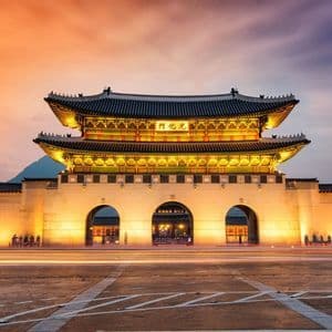 Ein traditionelles asiatisches Torhaus und seine Steinmauer sind in der Dämmerung hell erleuchtet, mit langen Lichtspuren von Fahrzeugen im Vordergrund.