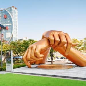 Eine große goldene Skulptur von zwei gekreuzten Händen in der 'Gangnam Style'-Tanzpose steht in einem Stadtpark vor einem modernen Wolkenkratzer.