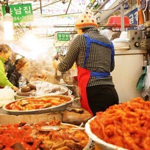 Un vendedor con gorro colorido y delantal cocina en un concurrido puesto de comida en un mercado interior, rodeado de grandes tazones humeantes de comida.