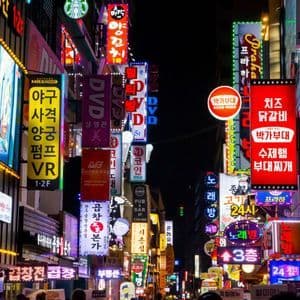 Una calle urbana estrecha de noche, brillantemente iluminada por una densa pared de coloridos letreros de neón, en su mayoría con escritura coreana.