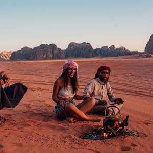 Una donna di un viaggio di gruppo WeRoad e due uomini siedono su dune di sabbia rosse, preparando il tè in un bollitore su un piccolo fuoco al tramonto.