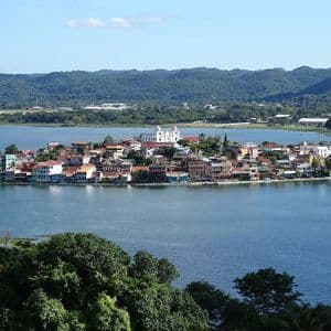 A daytime view of a town with colorful buildings on an island, surrounded by a blue lake and backed by green hills.