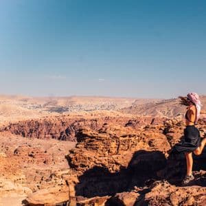 Una donna con un foulard posa su una gamba su uno sperone roccioso, affacciandosi su un vasto canyon desertico sotto un cielo azzurro e limpido.