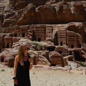 Una donna con capelli ricci e una fascia colorata sorride davanti a antiche tombe scavate in una parete rocciosa rossa.