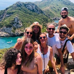 Un grupo de WeRoad sonríe para una selfie en un acantilado con vistas a una bahía turquesa y exuberantes colinas verdes.