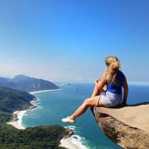 Una mujer se sienta al borde de un acantilado alto, mirando una costa montañosa con playas de arena y un océano azul.