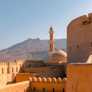 A mosque's golden dome and minaret rise above the sand-colored walls of a fortress, with distant mountains under a clear sky.