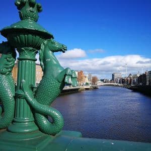 Un farol verde ornamentado con esculturas de hipocampos en un puente con vistas a un río que atraviesa una ciudad bajo un cielo azul.