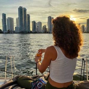 A woman with curly hair on a WeRoad group trip sits on a boat, watching the sun set behind a modern city skyline across the water.
