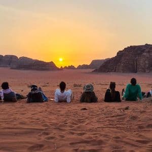 Un grupo de WeRoad en un viaje contempla el atardecer sobre la arena roja del desierto, con formaciones rocosas distantes al fondo.