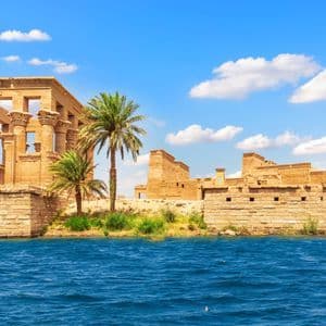 Un antico tempio di pietra con grandi colonne e palme sulla riva di un fiume sotto un cielo azzurro con nuvole.