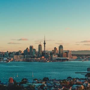 Eine Stadt-Skyline mit einem hohen Turm liegt bei Sonnenuntergang an einem Hafen, mit ruhigem blauem Wasser im Vordergrund.