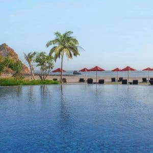 Una grande piscina a sfioro si affaccia su una spiaggia di sabbia con ombrelloni rossi, lettini e palme sotto un cielo limpido.