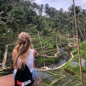 Una donna con una borsa WeRoad siede su un cornicione che si affaccia su una valle con risaie terrazzate verdi e alte palme.