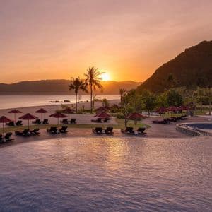 La piscina di un resort si affaccia su una spiaggia sabbiosa con lettini e ombrelloni rossi, mentre il sole tramonta sull'oceano e sulle colline.
