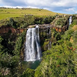 Una amplia cascada se precipita por un escarpado acantilado rocoso, rodeada de exuberante bosque verde y colinas onduladas bajo un cielo parcialmente nublado.
