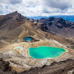 A WeRoad group trip hikes on a trail overlooking turquoise volcanic crater lakes in a mountainous landscape under a cloudy sky.