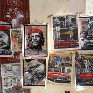 Eine Sammlung kubanischer Gemälde mit Che Guevara und Oldtimern hängt an einer Graffiti-Wand.