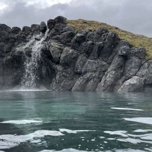 Ein kleiner Wasserfall stürzt über eine dunkle Felsformation in das dampfende Wasser einer natürlichen heißen Quelle.
