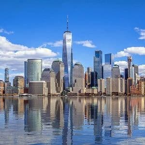A modern city skyline with tall skyscrapers reflecting in the calm water under a blue sky with scattered clouds.