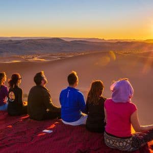 A WeRoad group trip sits on a blanket on a sand dune, watching the sunset over a vast desert landscape.