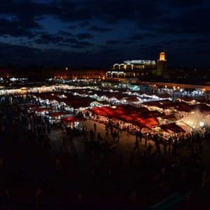 A bustling outdoor market square at night, filled with crowds of people among rows of illuminated stalls with red awnings.