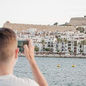 A person, seen from behind, takes a photo with a smartphone of a coastal town with white buildings and a hilltop fortress.
